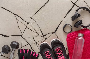 A pair of black and pink shoes sit beside weights and gloves on a cracked surface. A towel and water bottle are also visible. Headphones and a fitness band complete the workout setup.