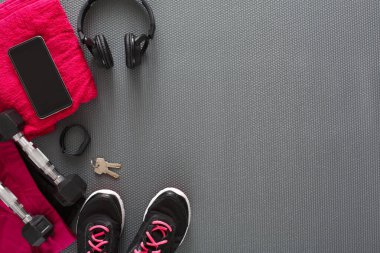 Bir insan gri bir spor paspası üzerinde egzersiz yapmaya hazırlanır. Pembe bir havlu, akıllı telefon, kulaklık ve anahtarlar yakınlarda. Bir çift siyah spor ayakkabı ve ağırlık da görülebilir..