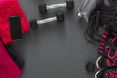 Black dumbbells sit on a gym mat next to a water bottle, a phone, workout gloves, a towel, and clothing. This setup is ready for an exercise session.
