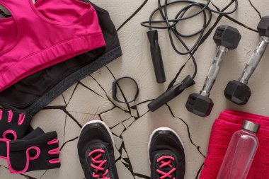 Bright workout gear is arranged on a cracked surface. There are dumbbells, a water bottle, and exercise tools nearby. This setting suggests preparation for a fitness routine or exercise session.