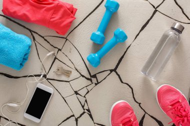 A workout space shows colorful clothes, dumbbells, a water bottle, headphones, and sneakers on a cracked floor. Personal items are neatly set for an exercise routine.
