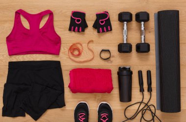 Bright workout clothing and fitness gear are arranged on a wooden floor. Items include dumbbells, a water bottle, a yoga mat, and a towel. This setup is ready for an exercise routine.