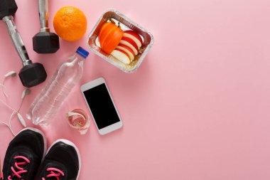 Dambıllar bir şişe su ve dilimlenmiş meyve konteynırının yanında durur. Yakınlarda kulaklıklı bir akıllı telefon var. Siyah spor ayakkabıları pembe yüzey üzerinde kullanıma hazır..