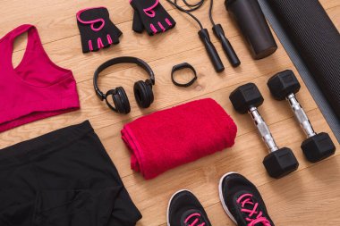 A variety of workout items are arranged on a wooden floor. The gear includes weights, a towel, gloves, a jump rope, a water bottle, and workout clothes. This setup is ready for exercise.