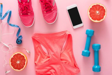 Bir çift pembe spor ayakkabı, bir telefon ve bir spor bluz pembe bir yüzeye kurulmuştur. Yakınlarda bir mezura, iki mavi dambıl ve yarım greyfurt sağlıklı bir egzersiz teması için yerleştiriliyor..