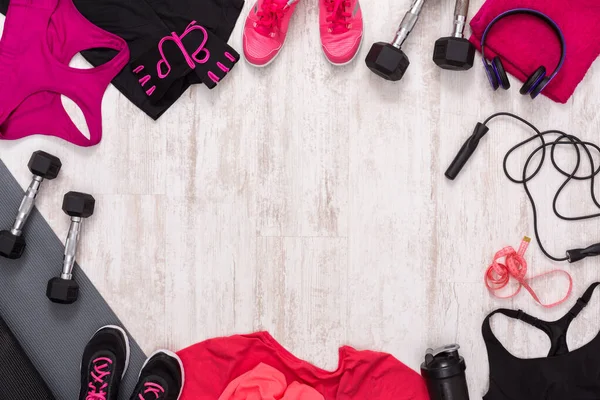 Various fitness items and clothing are spread out on the floor in a gym area. There are dumbbells, workout clothes, and shoes set for an exercise routine.