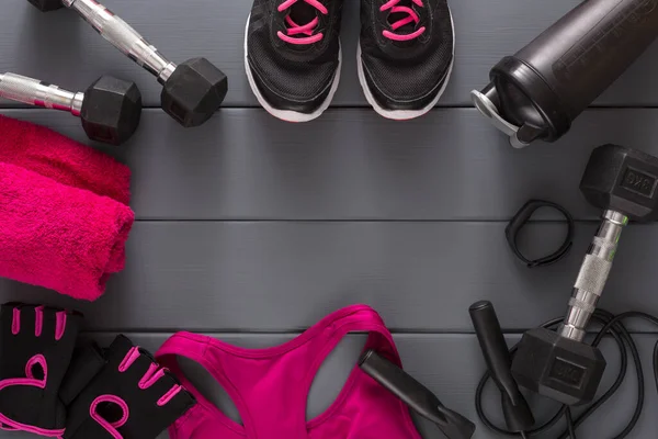 Various fitness items are laid out on a gray wooden floor. Items include dumbbells, a water bottle, workout gloves, and a towel. Exercise shoes are at the top center of the scene.