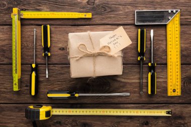 Kahverengi kağıda sarılı bir hediye ahşap bir yüzeye yerleştirilir. Hediyenin etrafında bantlar, tornavidalar ve Babalar Günü 'nüz kutlu olsun yazan bir not var..