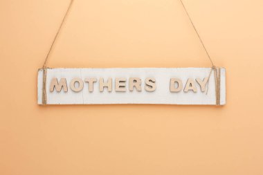 Açık turuncu renkli bir duvarda Anneler Günü yazılı bir tabela sergileniyor. Tabela ahşaptan yapılmış ve sicime asılmış, kırsal bir görünüm veriyor..