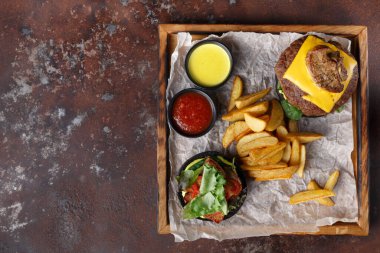 Peynirli bir hamburger, ahşap bir tepside patates kızartması ve küçük bir salatanın yanında durur. İki daldırma sosu da dahil. Restoran masasına benziyor..