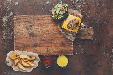 Ahşap bir tahta üzerinde marul ve peynir olan bir burger tutar. Yanlarında da ketçap ve hardal kaseleri vardır. Çıtır patatesler kenara konur. Bu yemek karanlık bir arka planda hazırlanmıştır..