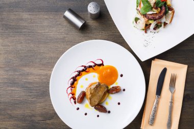 Bir restoranda ahşap bir masada iki tabak yemek sunulur. Bir tabakta et, sos ve süslemeler yer alıyor. Diğer yemek ise salata ve dekoratif malzemeler içeriyor..