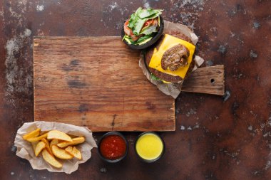Tahta kesme tahtasının üzerinde bir hamburger oturuyor. Hamburgerin üstünde peynir ve taze salata var. Yanında altın sarısı patates dilimleri ve yanında ketçap ve hardal kaseleri vardır..