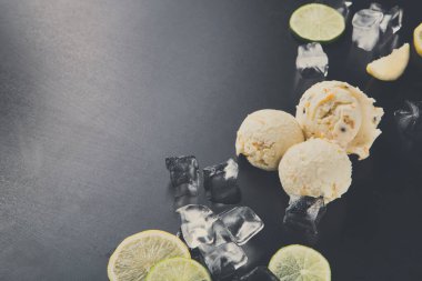 Üç kepçe dondurma koyu bir yüzeyde limon dilimleri ve buz kalıplarının yanında duruyor. Bu sahne, ferahlatıcı bir ikramla sıcak bir gün geçirmemizi öneriyor. Dondurma kremalı ve davetkar görünüyor..