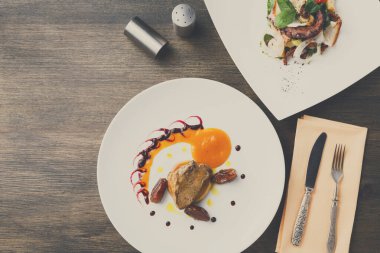 Tahta bir masada iki tabak yemek var. Bir tabakta soslu et ve hurma, diğerinde salata var. Gümüş çatal bıçak takımı tabağın yanında..