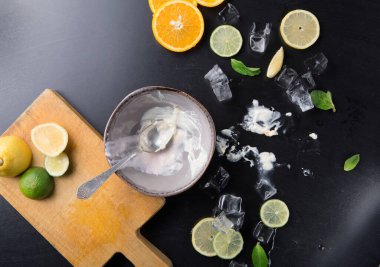 Mutfak tezgahında portakal, limon ve limon gibi taze meyve karışımları sergilenir. Yanında kaşıkla boş bir kase oturuyor. Buz küpleri ve nane yaprakları her yere dağılır..