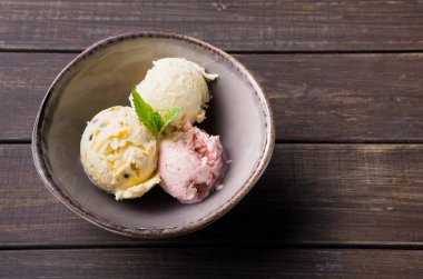 Kahverengi ahşap bir masanın üzerindeki bir kasede üç farklı dondurma çeşidi sunulur. Kepçeler beyaz, pembe ve sarı renkler içeriyor. Dekorasyon için üzerine bir nane yaprağı konur.