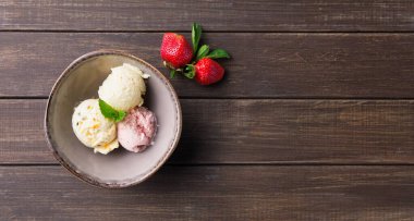Üç kepçe farklı dondurma, kahverengi bir kasede, ahşap bir masada parlak kırmızı çileklerin yanında oturur. Bu sahne neşeli bir yaz sürprizini yakalar..