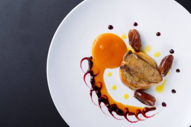 Renkli bir sosla servis edilen pişmiş tavuğun yer aldığı bir tabak ve hurmalar yuvarlak beyaz bir tabağa yerleştiriliyor. Tabak siyah bir zemine göre ayarlanmış, yemek düzenini vurguluyor..