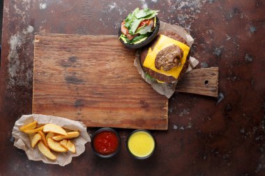 Bir hamburger eritilmiş peynir ve malzemelerle ahşap bir tahtaya yerleştirilir. Yanında çıtır patates kızartması ve taze salata var. İki küçük kase sos, yemeğe tat katar..