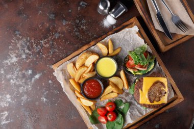 Yemek ahşap tepside servis edilir. Tabakta peynirli bir hamburger, taze marul ve domates var. Altın kızartmalar, soslar ve küçük domatesler var. Ayarlar gündelik..