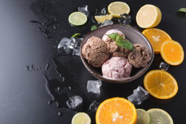 Kâselerde karanlık bir yüzeye yerleştirilmiş üç top dondurma var. Kasenin etrafında portakal, limon ve limon dilimleri var. Yüzeyde buz küpleri var..