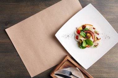 Restorandaki bir masanın üzerine renkli bir tabak konur. Tabakta deniz ürünleri ve sebze gibi çeşitli malzemeler yer alıyor. Yemek salonunda basit dekorlu ahşap bir masa var..