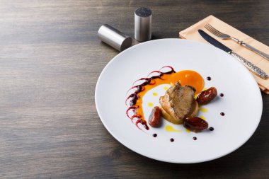 Tabakta, ahşap bir masanın üzerinde meyve püresi ile süslenmiş sosun yanında et sergileniyor. Gümüş çatal bıçak takımı ve peçete yakında. Yemek, bir restorandaki yemek deneyiminin bir parçası..