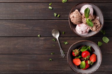 İki kase ahşap bir masanın üzerinde. Bir kasede farklı dondurma çeşitleri var. Diğer kasede taze çilek var. Nane yaprakları kaselerin etrafına saçılmış..