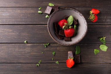 Taze çilekler, siyah çikolata parçaları ve ahşap bir masanın üzerindeki nane yapraklarının yanında bir kasede otururlar. Düzenleme, atıştırmalık ya da tatlı için basit ve eğlenceli bir ikram sunuyor..