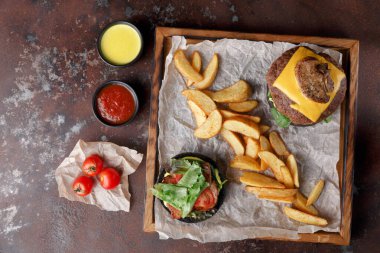 Tahta tabakta bir hamburger, kızarmış patates, küçük bir kase salata ve iki sos var. Taze domatesler yakına yerleştirilir ve basit bir yemek düzenine dönüştürülür..