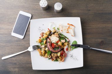 Bir tabakta sebze ve patatesle karıştırılmış ahtapot salatası var. Yakınlarda bir akıllı telefon var. Tuz ve biberlikler sofra düzenini tamamlar. Yemek bir lokantada yenmeye hazır..