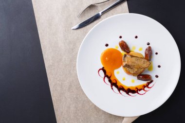 Bir parça et, sos ve süslemeler de dahil olmak üzere çeşitli elementlerle beyaz bir tabakta düzenlenir. Çatal bıçak takımı yakında ve masa koyu ve açık renkli..