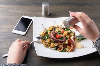 Bir el sebzeli deniz ürünleri makarnasına tuz ekliyor. Masanın yanında bir akıllı telefon var. Yemek taze ve renkli görünüyor. Bu sahne modern bir yemek salonunda geçiyor..