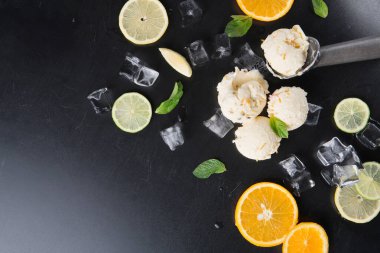 Dondurma parçaları, siyah bir yüzeydeki buz küpleri arasında dinleniyor. Dondurmanın etrafını limon ve portakal dilimleri sarar ve taze nane yaprakları olay yerine renk katar. Ayarlar tatlı zamanı olduğunu gösteriyor..