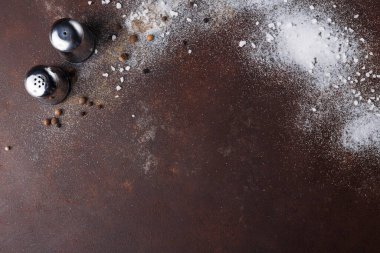 İki metal tuz ve biber karıştırıcı kahverengi bir yüzeye dağılmış tuz ve biberle saçılmıştır. Sahne, çeşitli baharatlarla dolu kırsal bir mutfak ortamını gösteriyor..