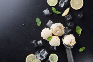 Limon dilimleri, nane yaprakları ve buz küpleriyle çevrili siyah bir masada bir kepçe dondurma duruyor. Sahne, sıcak bir günde tadını çıkarmaya hazır ferahlatıcı bir tatlıyı gösteriyor..