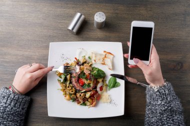 Renkli sebze ve et içeren bir yemeği yemek için bir çatal kullanır. Öte yandan bir telefon tutulur. Sofrada bir tuzluk ve biraz ekmek var..