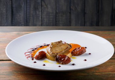 Beyaz bir tabakta bir tabak pişmiş et sunulur. Sos, garnitür ve vişneli domates de var. Ahşap masa akşam yemeği sırasında restoran atmosferine sıcaklık katar..