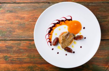 Özenle hazırlanmış bir tabakta bir parça et, parlak bir portakal sosu ve birkaç küçük parça eşya görünüyor. Renkli soslar yemeğin etrafında sanatsal bir şekil oluşturur. Masa ahşap..