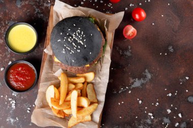 Siyah burger yanında patates kızartması olan kahverengi bir kağıt parçasına oturur. Hamburgerin yanında soslu iki küçük kase var. Taze kiraz domatesleri tabağın etrafına saçılmış..