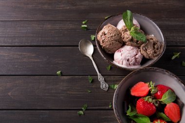 İki kase ahşap bir masaya yerleştirilir, bir kase farklı dondurma çeşitleri içerir, ve diğeri taze çilek içerir. Kâselerin yanında bir kaşık yatıyor..