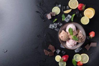 Bir kâsede bir kepçe dondurma var. Kasenin etrafında limon ve limon dilimleri, çilekler, çikolata parçaları ve koyu bir yüzeyde buz küpleri bulunuyor. Bu sahne çekici ve taze..