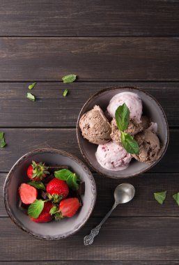 İki kase ahşap bir masada oturur. Bir kasede renkli dondurma kepçeleri, diğerinde taze çilekler var. Yeşil nane yaprakları biraz renk katar.