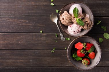 İki kase ahşap bir masanın üzerinde. Bir kasede farklı dondurma çeşitleri var. Diğer kasede taze çilek var. Nane yaprakları kaselerin etrafına saçılmış..
