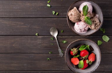İki kase ahşap bir masanın üzerinde. Bir kasede farklı dondurma çeşitleri, diğerinde taze çilek ve çikolata var. Nane yaprakları etrafa saçılmış..