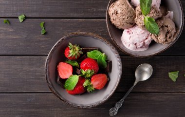 Bir kase taze çilek ve çikolata parçalarının yanında bir kase karışık dondurma içerir. Nane yaprakları tatlıyı süslüyor. Masanın üzerinde bir kaşık duruyor..