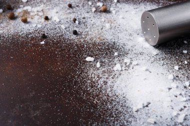 Tuz, dağınık karabiberlerin yanında, koyu ahşap bir yüzeyde bulunur. Yakınlarda yemek pişirmenin sonuçlarını gösteren bir metal çalkalayıcı var. Yemek hazırlama için gerekli malzemeler mevcut..