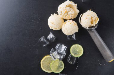 Siyah bir yüzeye üç kepçe limonlu dondurma konur. Dondurmanın etrafında dilimlenmiş limonlarla birlikte buz parçaları var. Sahne taze ve davetkar görünüyor..