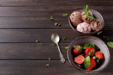 İki kase dondurma ve ahşap bir masanın üzerine yerleştirilmiş taze çilekler. Kâselerin yanında bir kaşık yatıyor. Nane yaprakları sahneye biraz renk katar ve sıradan bir tatlı anı yaratır..